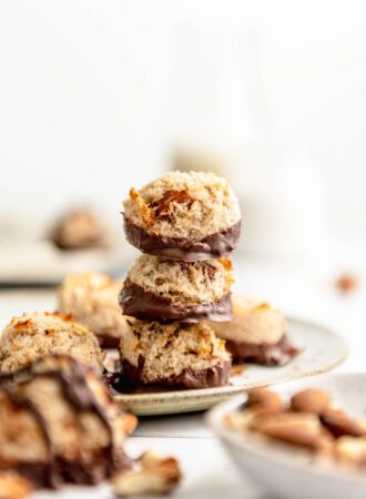 Stack of 3 coconut almond macaroon haystack cookies dipped in chocolate on a plate with more macaroons scattered around in the background and foreground.