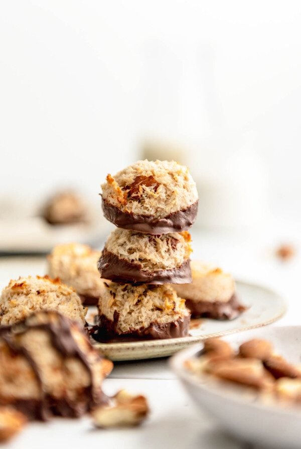 Stack of 3 coconut almond macaroon haystack cookies dipped in chocolate on a plate with more macaroons scattered around in the background and foreground.