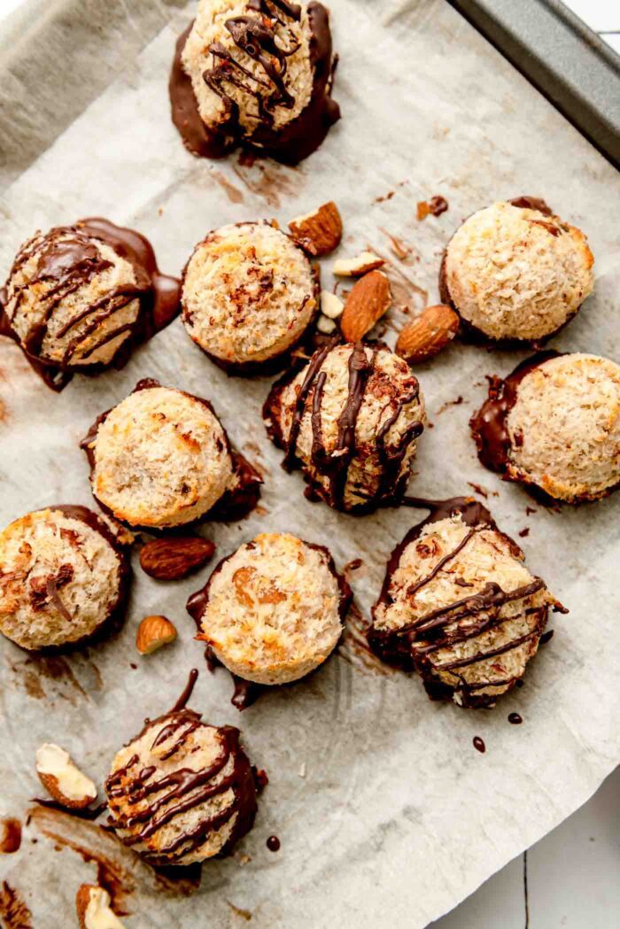 Overhead view of a number of almond macaroons dipped in chocolate and drizzled with chocolate on a baking tray lined with parchment paper.