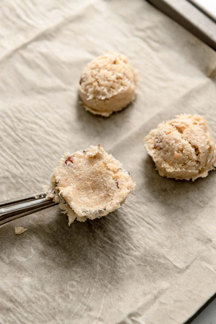 Almond macaroon cookie dough pressed into a small cookie scoop on a baking tray with two almond macaroons beside it.