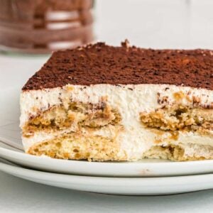 Square of tiramisu cake on a plate. You can see the layers of ladyfingers, cream filling and cocoa powder on top.