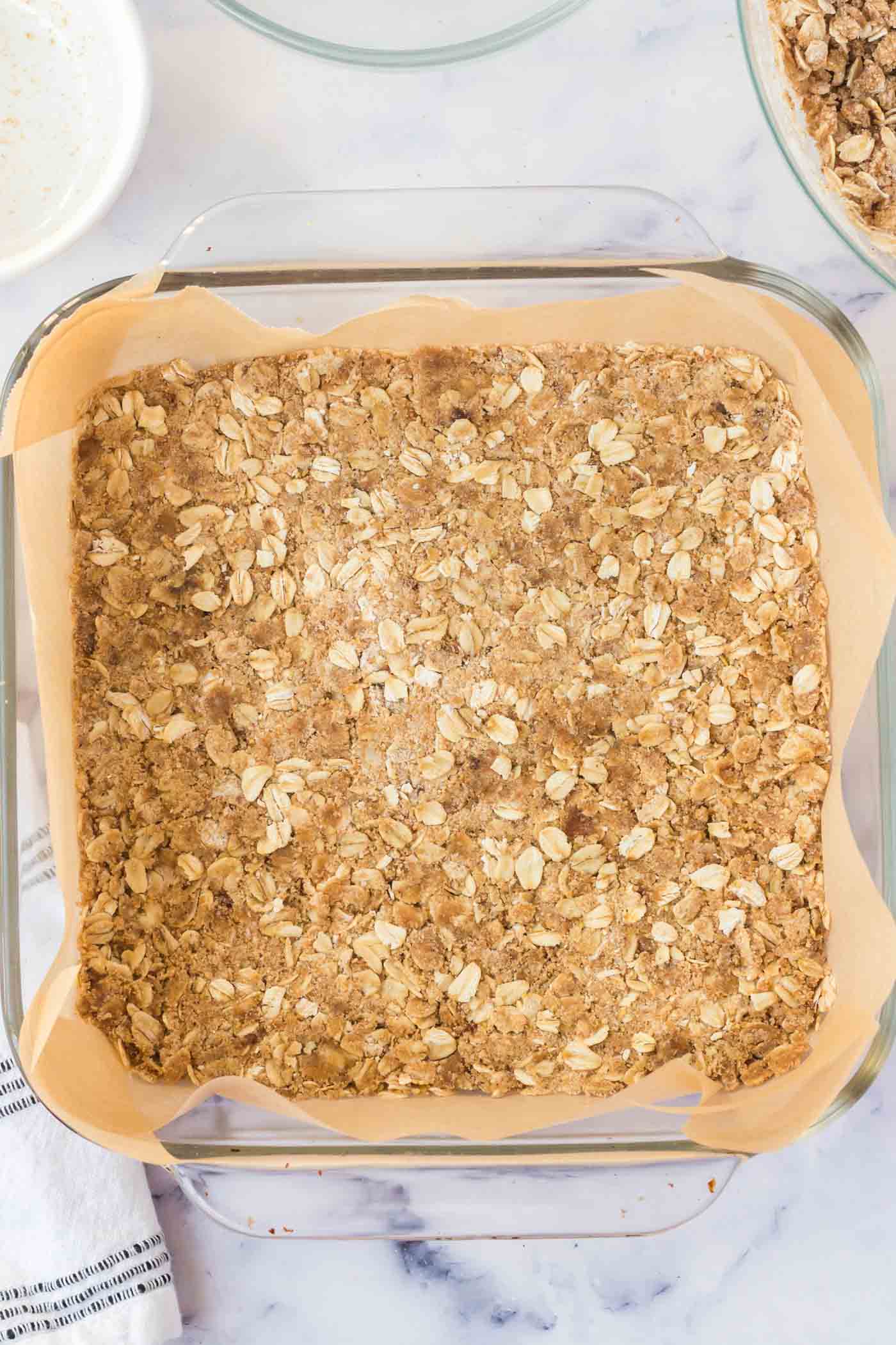 An oatmeal crust pressed into a square glass baking dish lined with parchment paper.