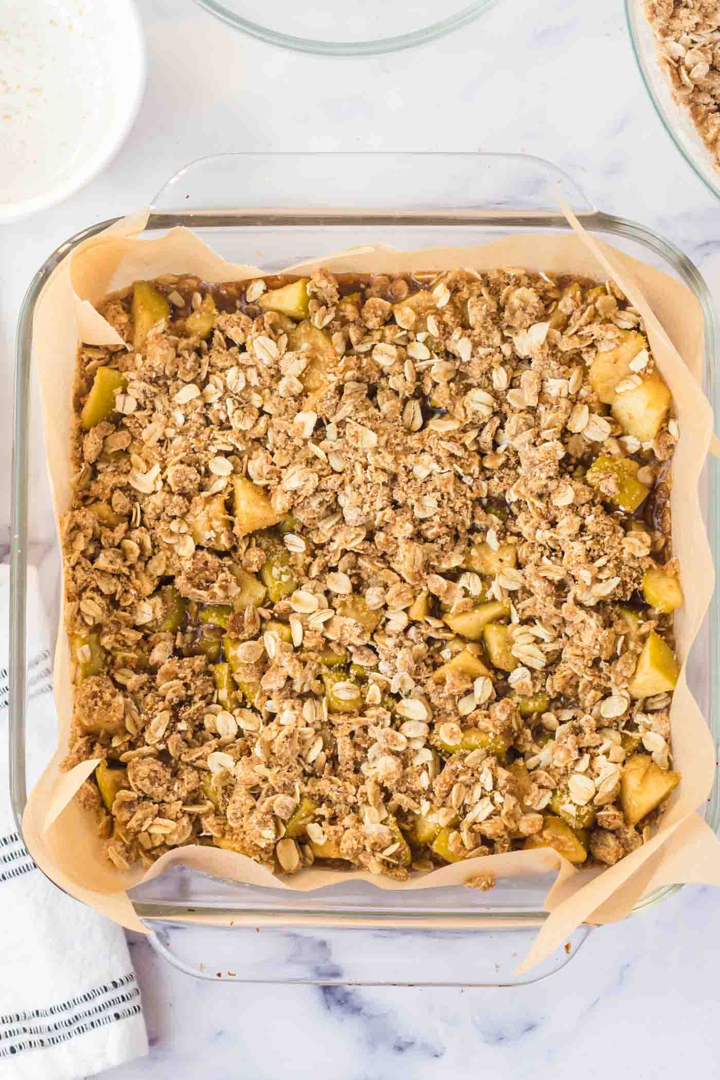 Oatmeal streusel topping sprinkled over apple crumble bars in a square glass baking dish.