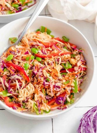 A bowl of ramen noodle salad with shredded red cabbage, green cabbage, carrot, edamame, cashews, green onions and red bell pepper. There's a fork resting in bowl and a chunk of red cabbage just out of the frame in the bottom right corner.