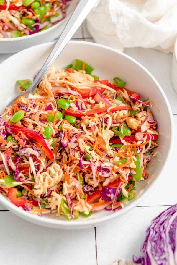 A bowl of ramen noodle salad with shredded red cabbage, green cabbage, carrot, edamame, cashews, green onions and red bell pepper. There's a fork resting in bowl and a chunk of red cabbage just out of the frame in the bottom right corner.