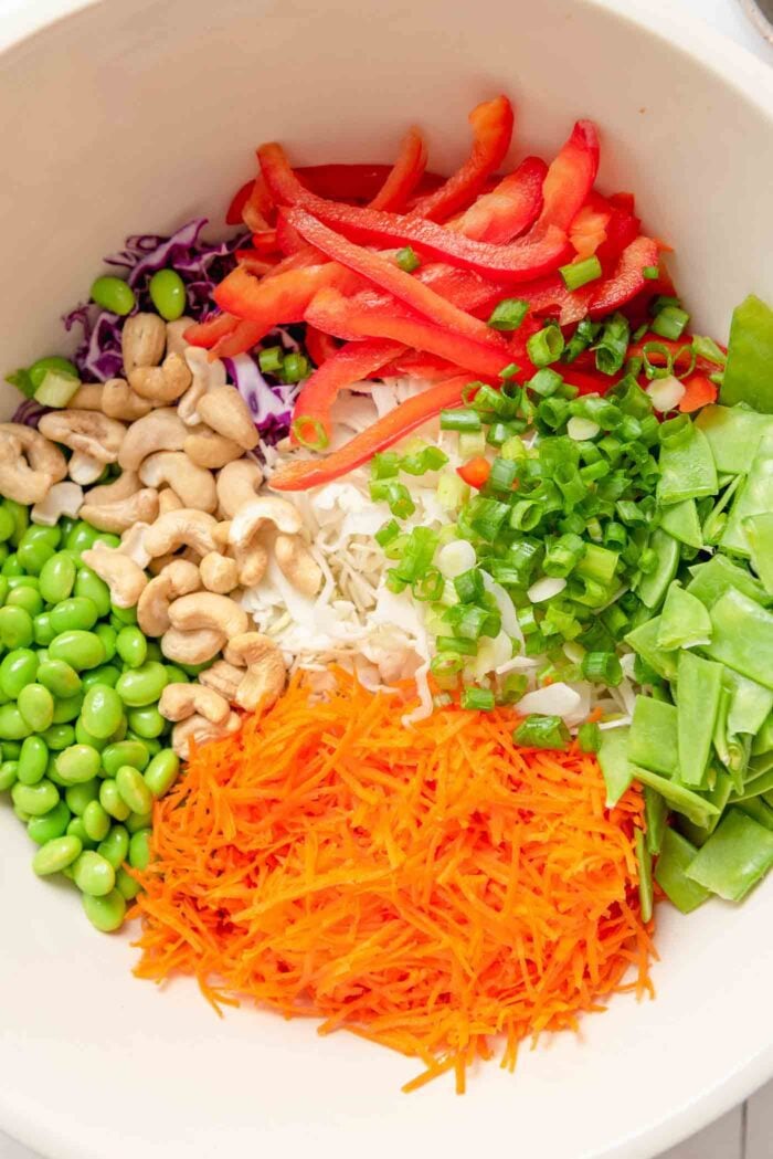 Grated carrot, chopped snow peas, bell pepper, cashews, edamame, shredded red cabbage and green cabbage in a large ceramic mixing bowl.