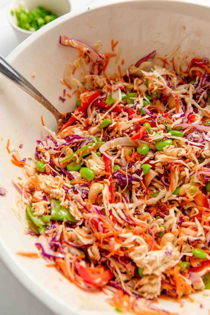 Ramen noodle salad with red cabbage, green cabbage, cashews, edamame and carrot in a large mixing bowl with a metal serving spoon resting in the bowl.