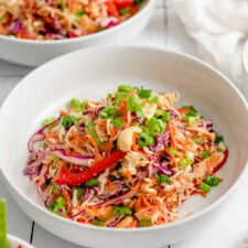 A bowl of crunchy Asian ramen noodle salad with cashews, edamame, bright coloured vegetables like cabbage, carrot, green onion and bell pepper. There is another bowl of salad in the background.