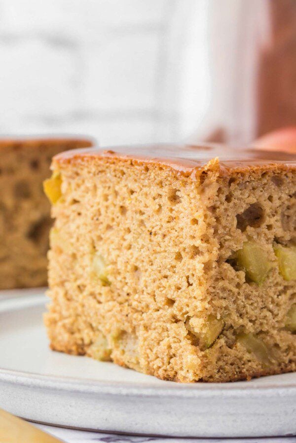 Close up of a large square of apple spice cake from an angled view on a small plate. You can see bits of apple in the cake and it's topped with a thin layer of caramel.