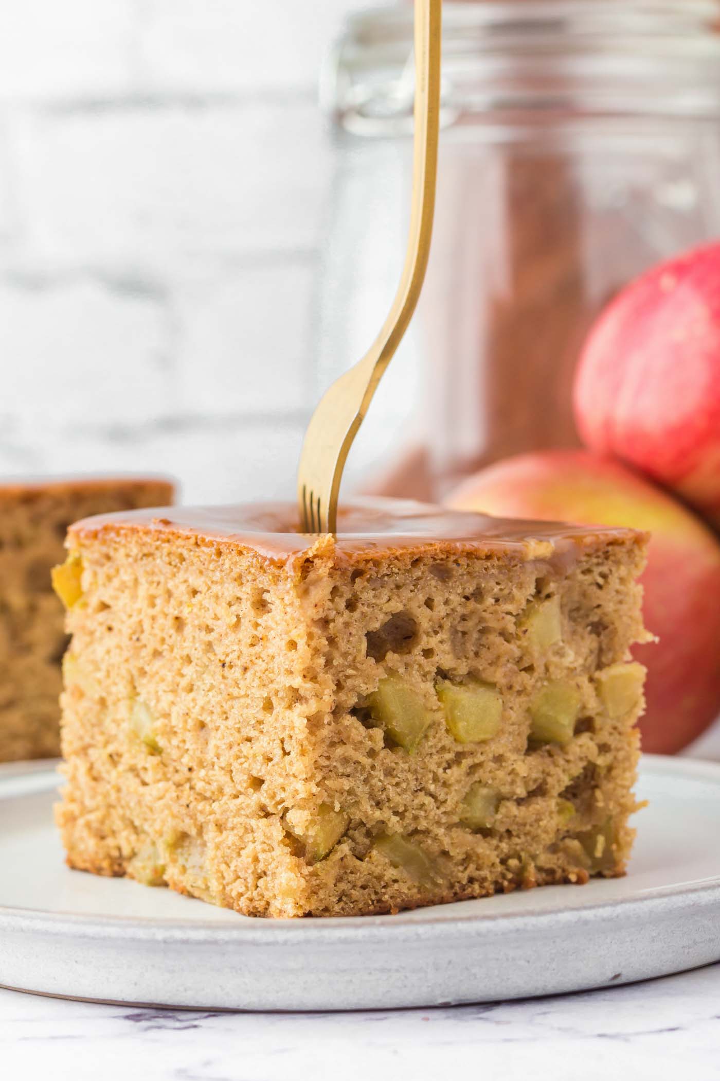 A large square of vegan apple cake on a plate. The cake is topped with a caramel glaze, there's a fork stuck in the cake and you can see some pieces of diced apple in the cake.