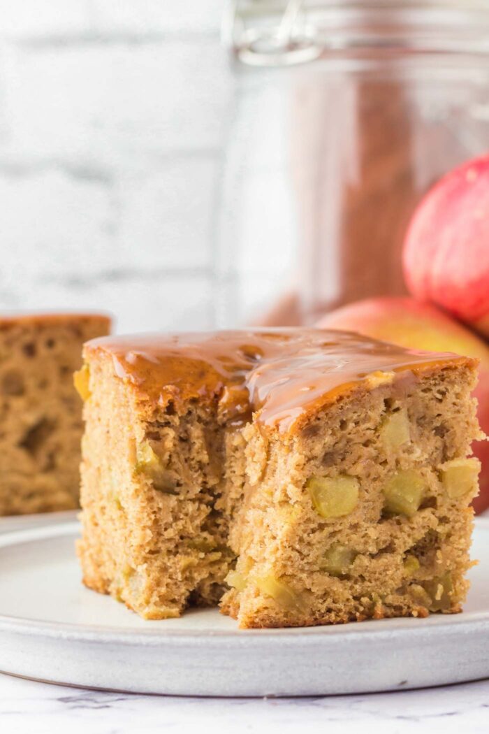 A large square of caramel apple cake with a forkful taken from it on a plate. There are pieces of diced apple in the cake.