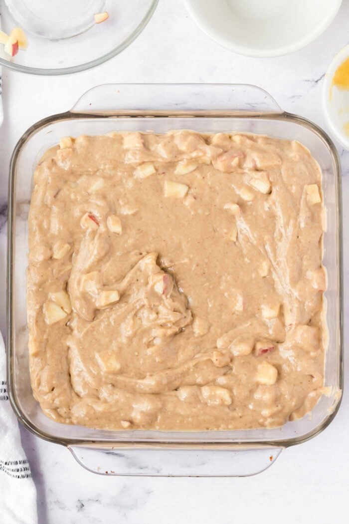 Raw apple cake batter with pieces of apple in it in a square glass baking dish.