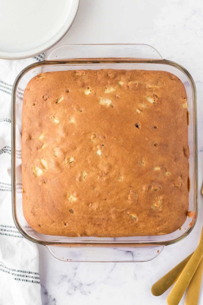 Overhead view of a baked apple cake in a square glass baking dish. You can see pieces of diced apple in the cake and the cake is a light golden brown colour.