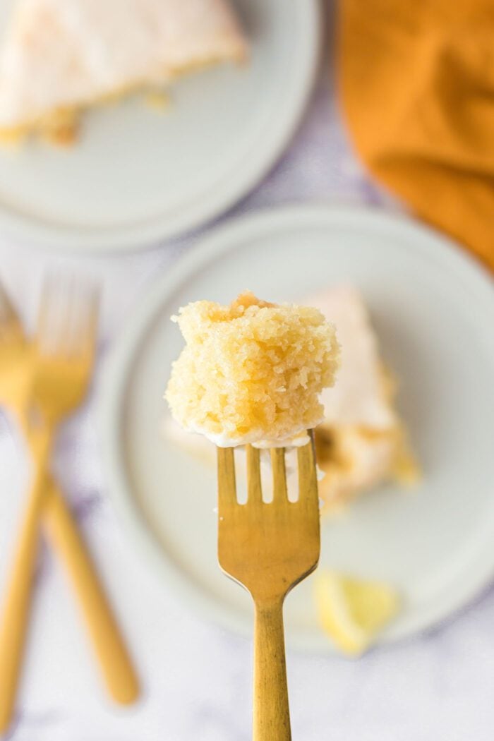 A small piece of olive oil cake with icing on the end of a fork held over a plate with a slice of cake on it.