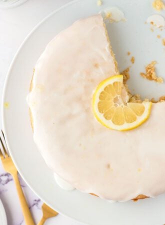 Overhead view of an olive oil cake topped with a powdered sugar icing on a cake stand. There's a slice of lemon decorating the middle of the cake and a slice out of the cake.