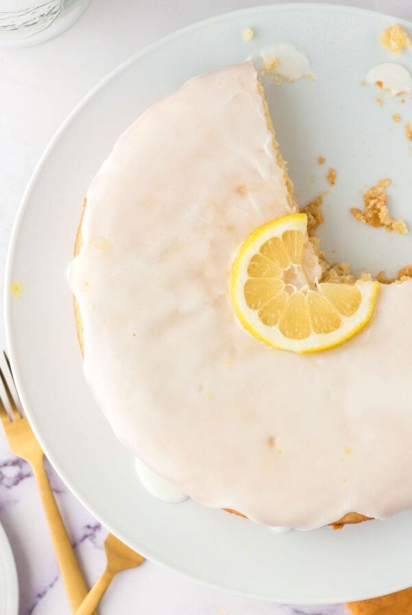 Overhead view of an olive oil cake topped with a powdered sugar icing on a cake stand. There's a slice of lemon decorating the middle of the cake and a slice out of the cake.