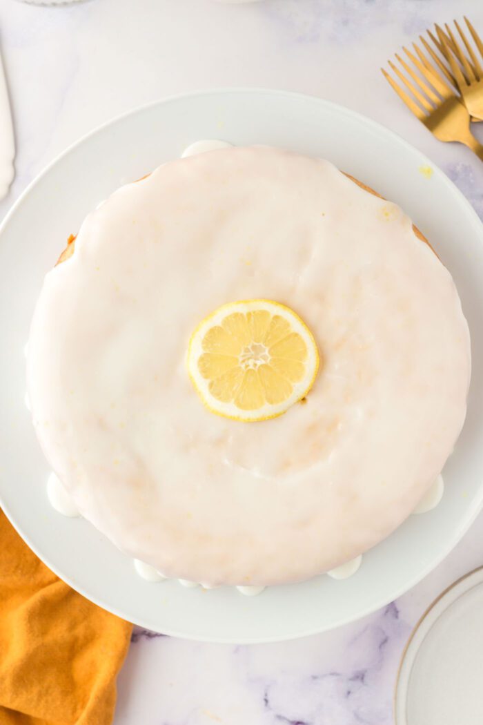 Overhead view of an olive oil cake topped with a powdered sugar icing on a cake stand. There's a slice of lemon decorating the middle of the cake.