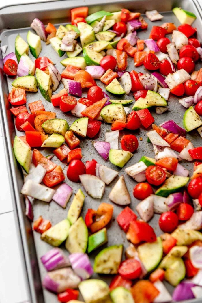 Chopped red onion, bell pepper, zucchini and cherry tomato tossed with herbs and oil on a baking pan.
