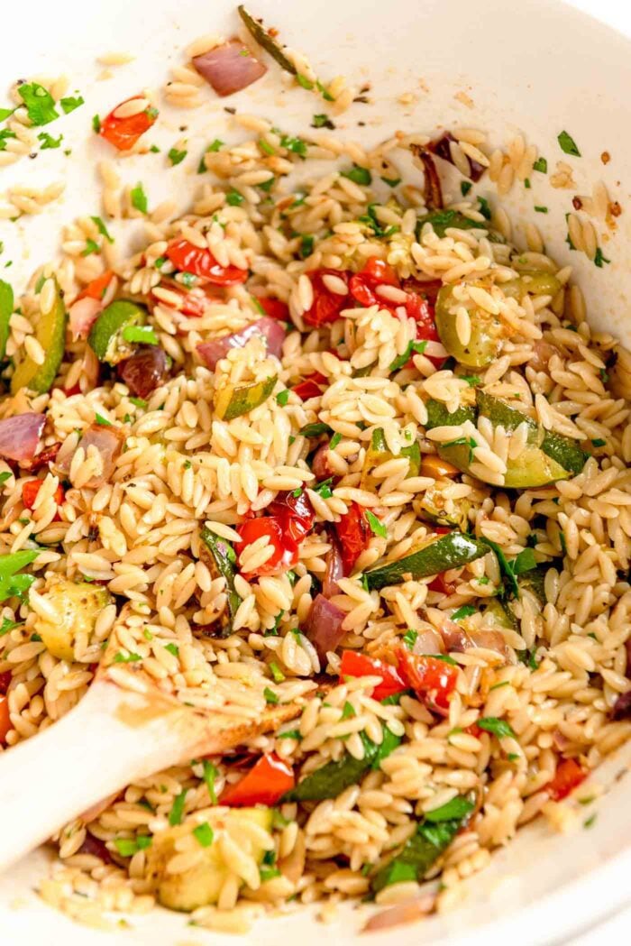 Orzo mixed with roasted red onion, tomato, zucchini and bell pepper in a large bowl with a wooden spoon resting in it.