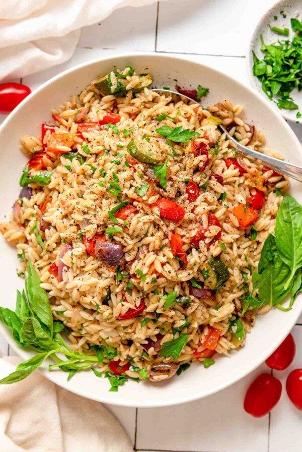 Overhead view of orzo with roasted vegetables garnished with a few pieces of fresh basil. There are a few cherry tomatoes beside the bowl and a small dish of chopped fresh parsley.