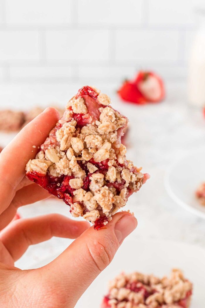 A hand holding a strawberry crumble bar with a bite taken out of it. There are a few fresh strawberries in the background and a plate with more bars on it.