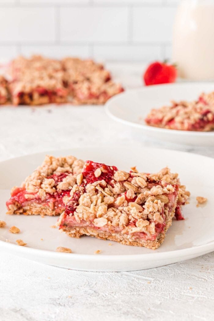 Two strawberry crumble bars on a plate. There's another plate of 2 oat bars in the background as well as the whole pan of bars on a piece of parchment paper.