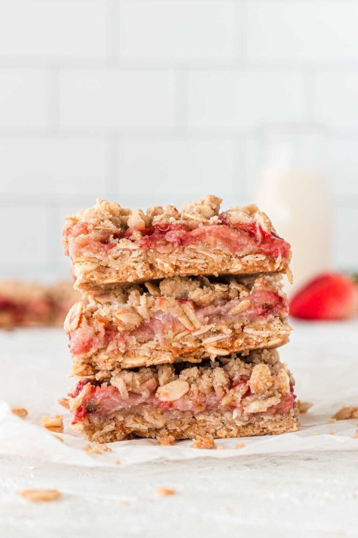 A stack of 3 strawberry oatmeal crumble bars on a small piece of parchment paper against a subway tile background. There is a fresh strawberry and a jar of milk in the backround.