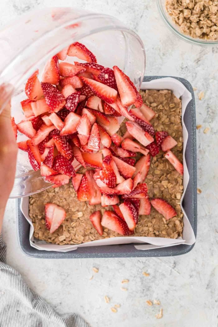 Dumping diced strawberries from a glass bowl onto an oatmeal crust in a square baking lined with parchment paper.