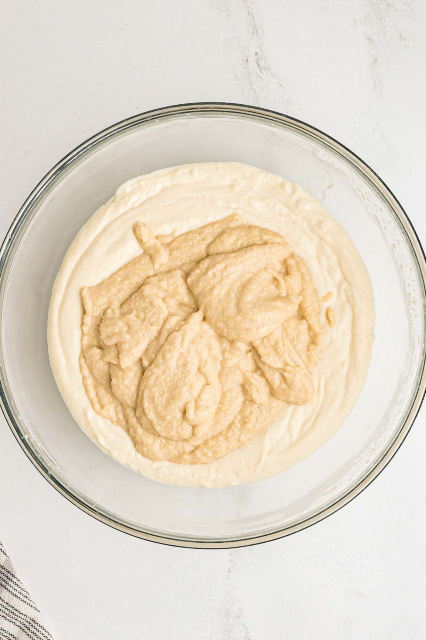 A creamy blended cashew mixture poured over a cream cheese-like mixture in a glass mixing bowl. Each cream is a slightly different shade of beige.