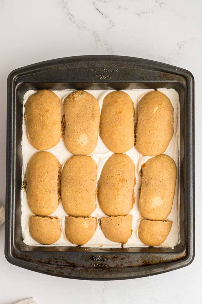 A layer of golden brown ladyfinger cookies on a layer of creamy marscapone in a square baking pan.