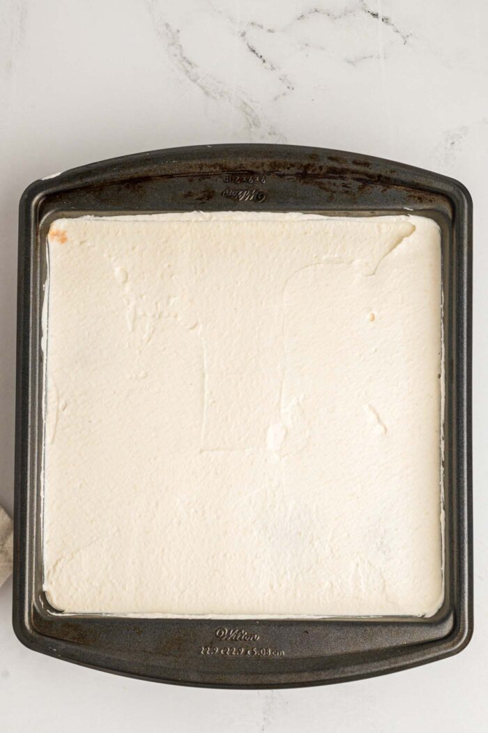 Marscapone cream cheese spread in an even layer in a square baking pan of tiramisu cake.