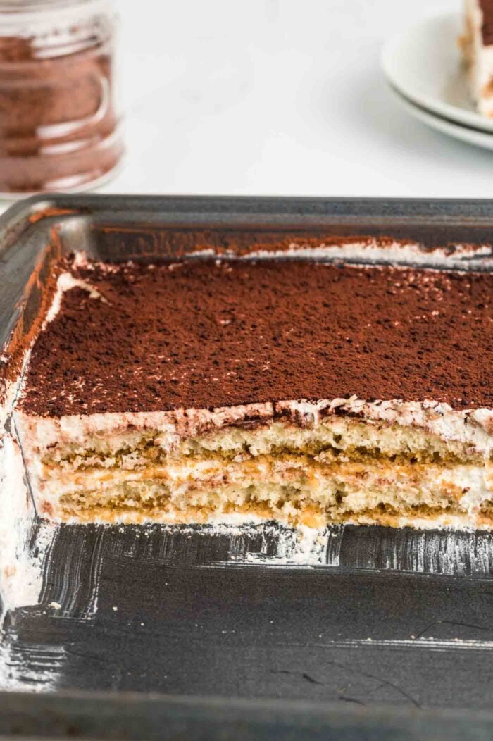 Half of a vegan tiramisu in a pan so you can see the layers of ladyfingers and cream filling.