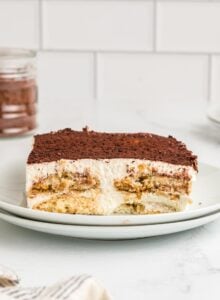 A square serving of tiramisu cake on a plate. You can see the layers of ladyfingers, marscapone cream filling and cocoa powder on top.