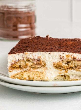 Close up of a square of tiramisu cake on a plate. You can see the layers of ladyfingers, cream filling and cocoa powder on top.