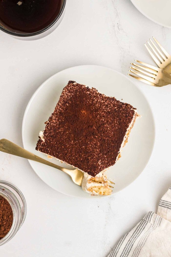 Overhead view of a large square of tiramisu topped with cocoa powder on a small plate. A fork with a piece of cake on it rests on the plate.