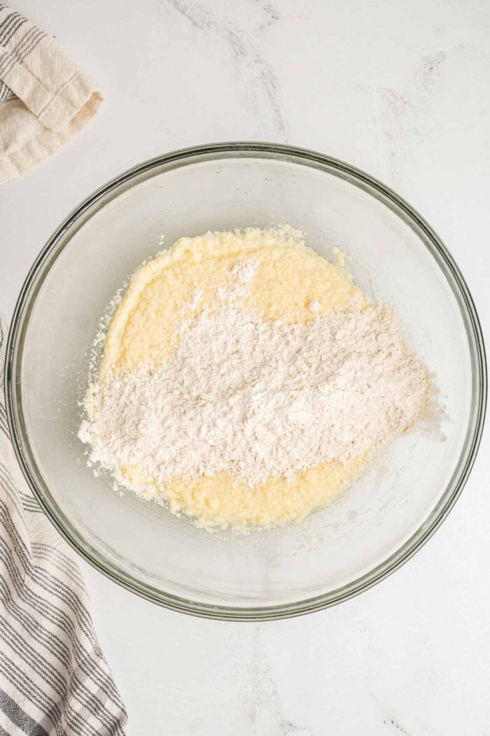 A creamed butter and sugar mixture in a glass mixing bowl with some flour added on top of it, ready to be mixed together.