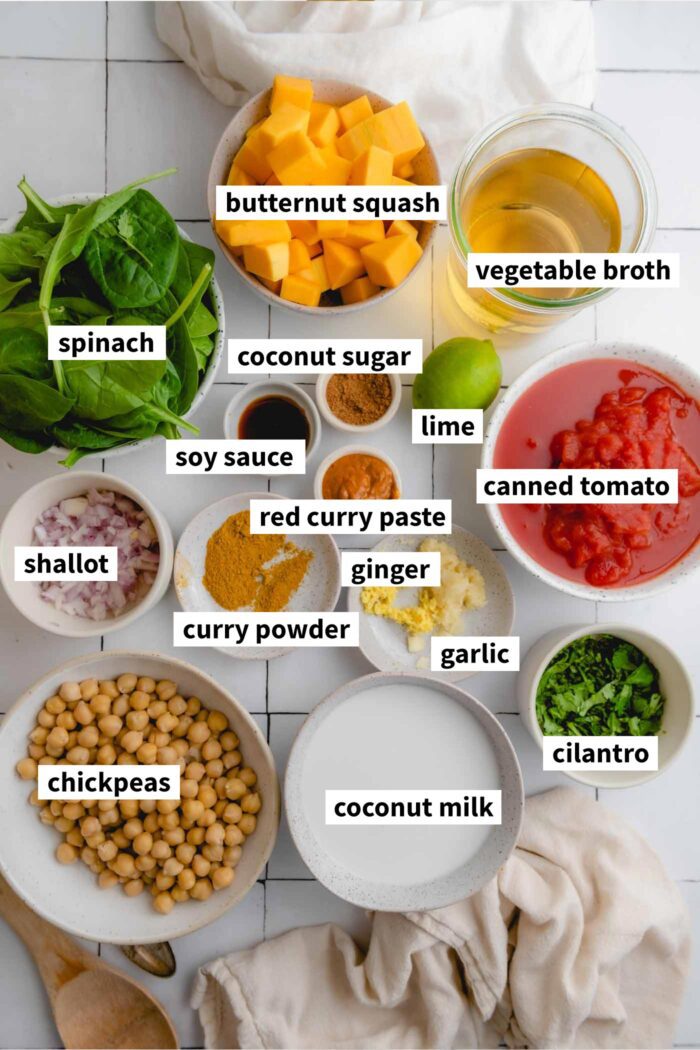 All the ingredients for making a butternut squash curry recipe with spinach, chickpeas, coconut milk and tomato. Each ingredient is labelled with text.