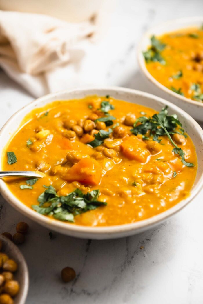 A bowl of sweet potato lentil soup topped with crispy chickpeas and cilantro. A second bowl can be seen in the background.