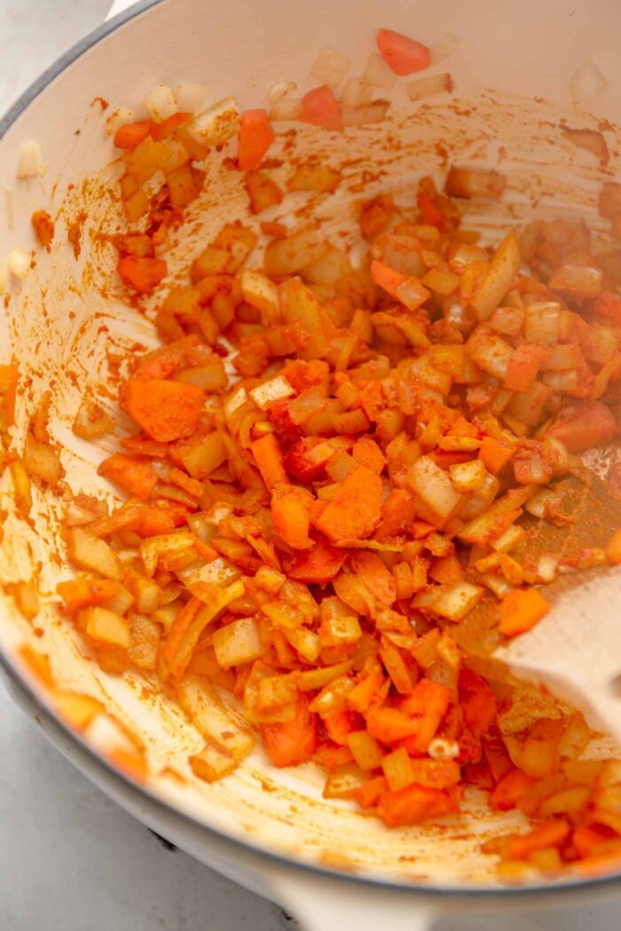 Chopped onion and carrot cooking in a spice mixture in a large soup pot with a wooden spoon.