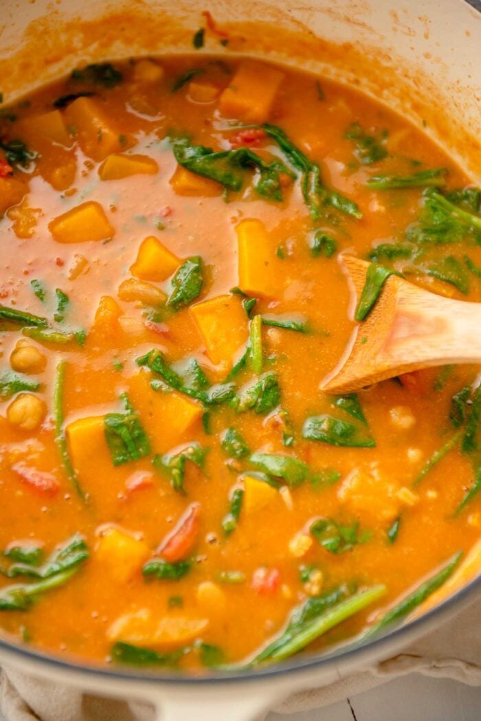 A large pot of butternut squash curry with spinach and chickpeas. A wooden spoon rests in the pot.
