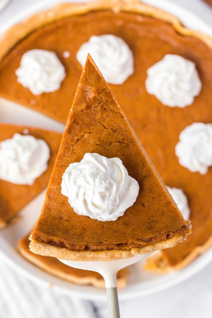 A slice of sweet potato pie topped with a dollop of whipped cream on a metal spatula held over the rest of the pie in a pie dish.