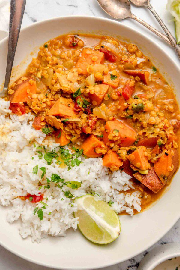 Sweet potato and red lentil curry in a bowl with rice and a wedge of lime. A spoon rests in the bowl and there are two more small spoons beside the bowl on the counter.