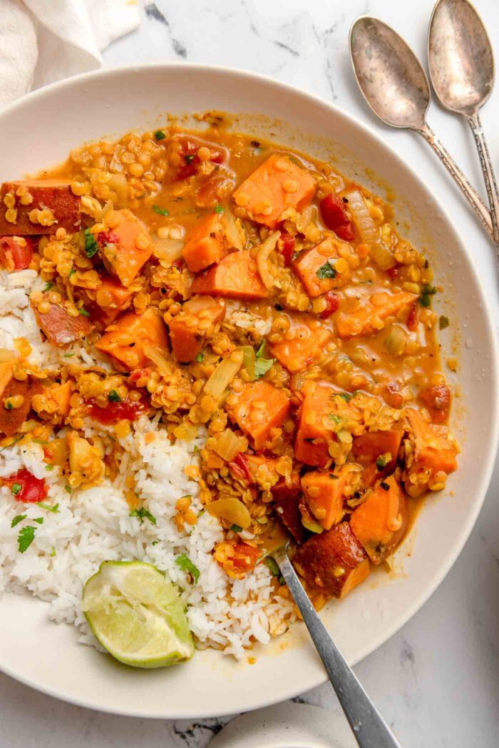 Overhead view of a sweet potato and red lentil curry in a bowl with rice and a wedge of lime. A spoon rests in the bowl and there are two more small spoons beside the bowl on the counter.