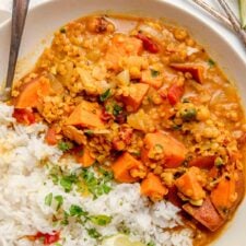 Overhead view of a sweet potato and red lentil curry in a bowl with rice and a wedge of lime. A spoon rests in the bowl and there are two more small spoons beside the bowl on the counter.