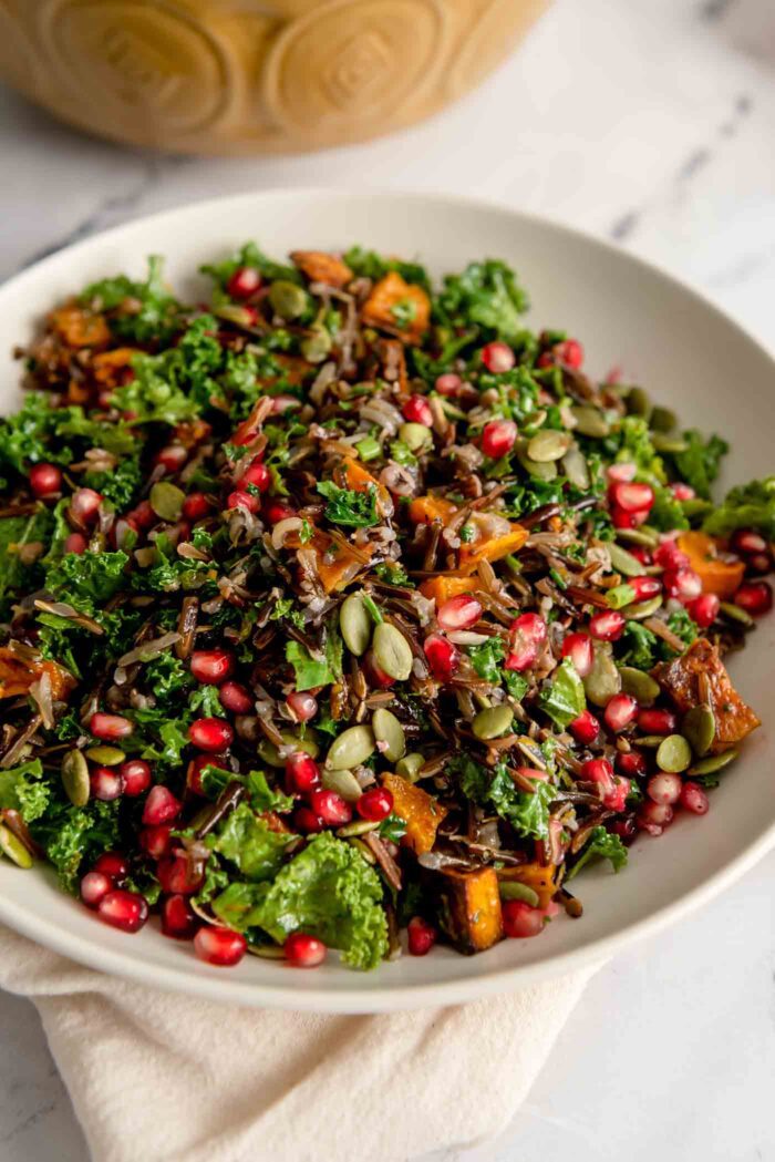 A bowl of wild rice salad with pomegranate arils, squash, kale, pumpkin seeds and green onion.