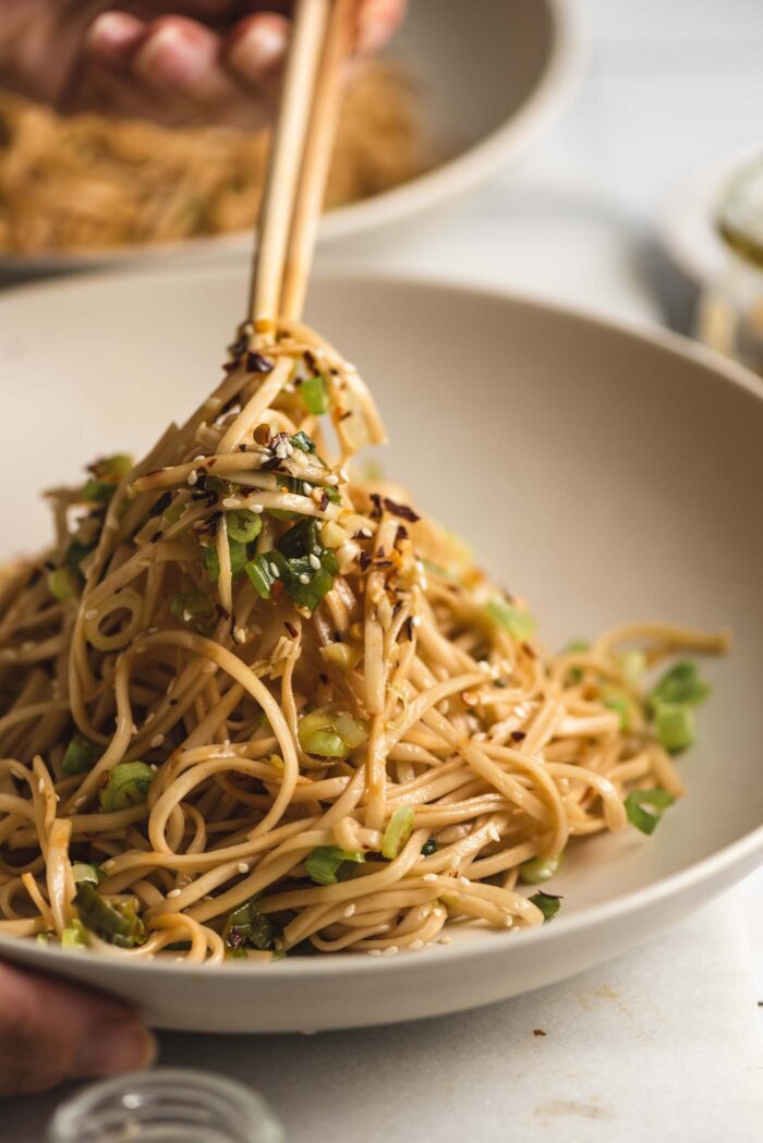 Chopsticks lifting garlic chili oil noodles with scallions and green onions from a bowl.
