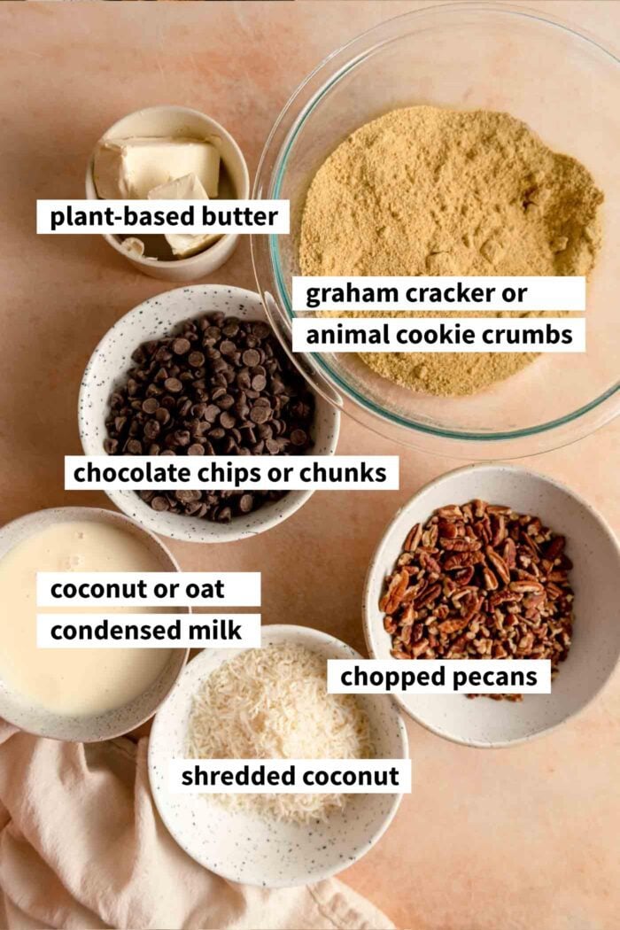 All the ingredients needed for making seven layer cookies bars. Each ingredient is in a bowl and labelled with text overlay.