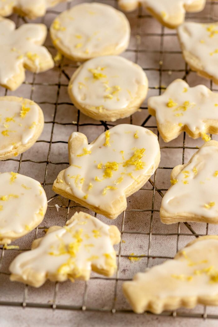 Glazed lemon shortbread cookies sprinkled with lemon zest on a cooling rack. The cookies are cut out in Christmas themed shapes like mittens and trees.