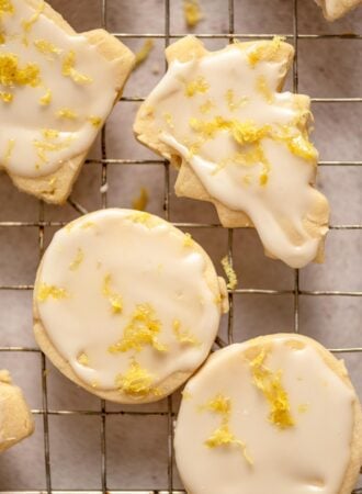 A tree-shaped and two round lemon shortbread cookies iced with lemon glaze and sprinkled with lemon zest on a cooling rack.