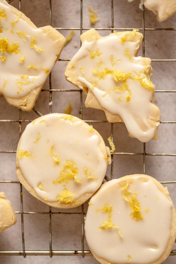 A tree-shaped and two round lemon shortbread cookies iced with lemon glaze and sprinkled with lemon zest on a cooling rack.